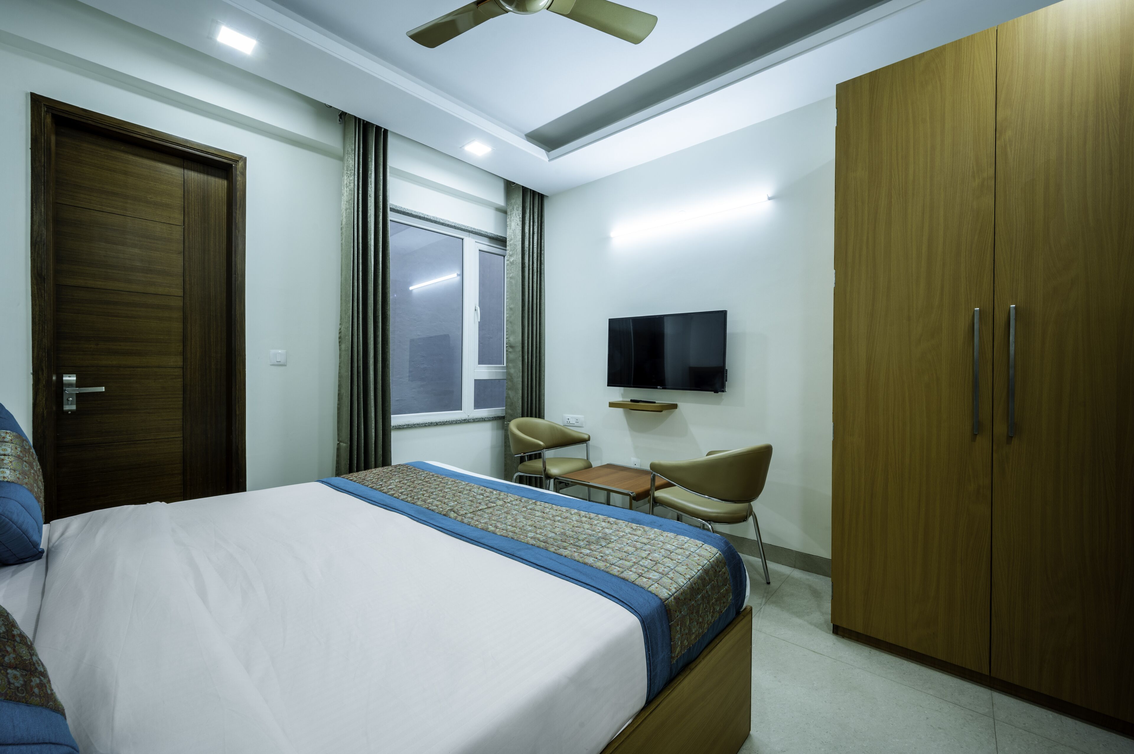 A hotel room with a bed, a desk, two chairs, a wardrobe, a television, and a ceiling fan.