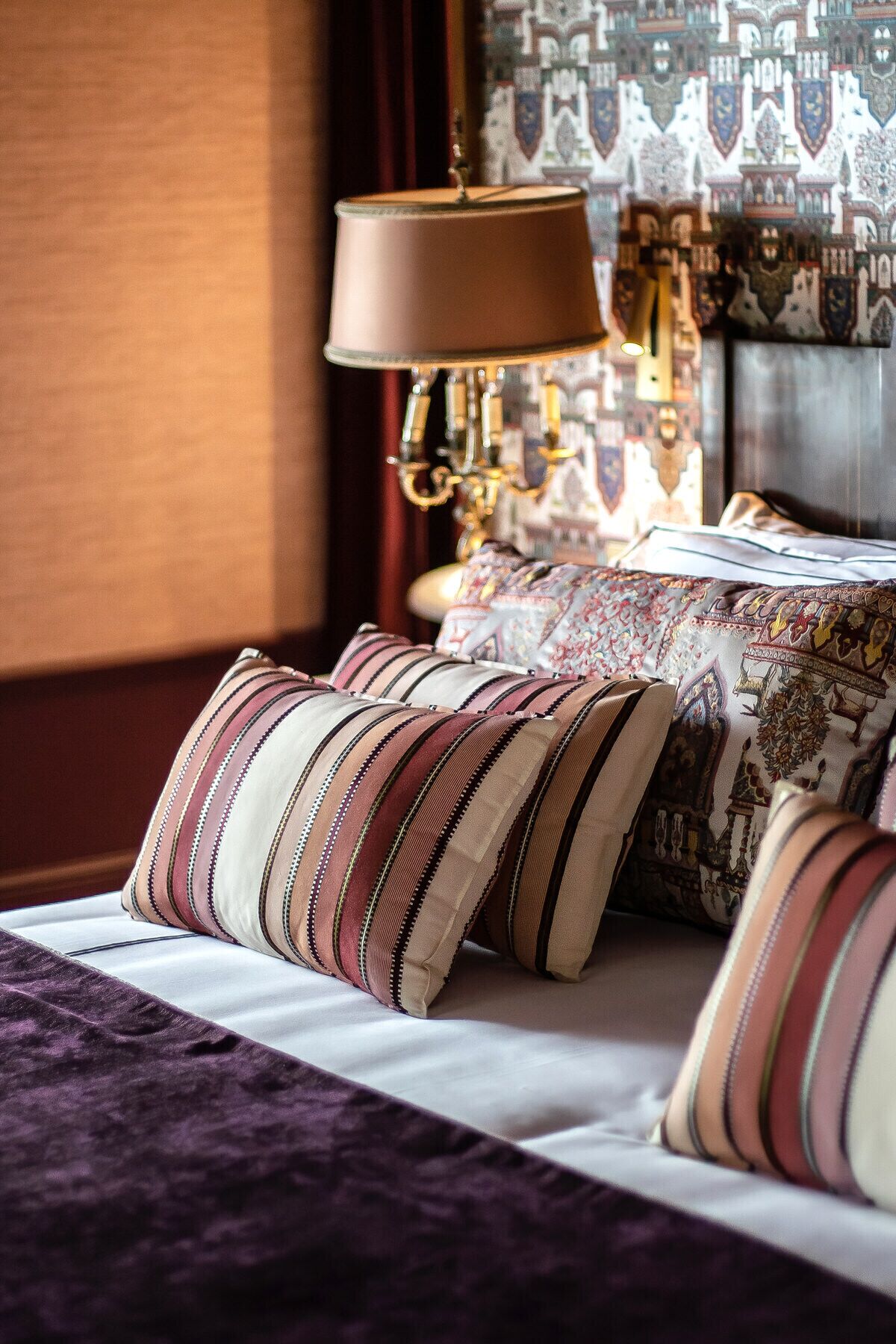 Una cama con almohadillas a rayas y una colcha estampada, una lámpara con pantalla color beige y una pared decorada con un diseño.