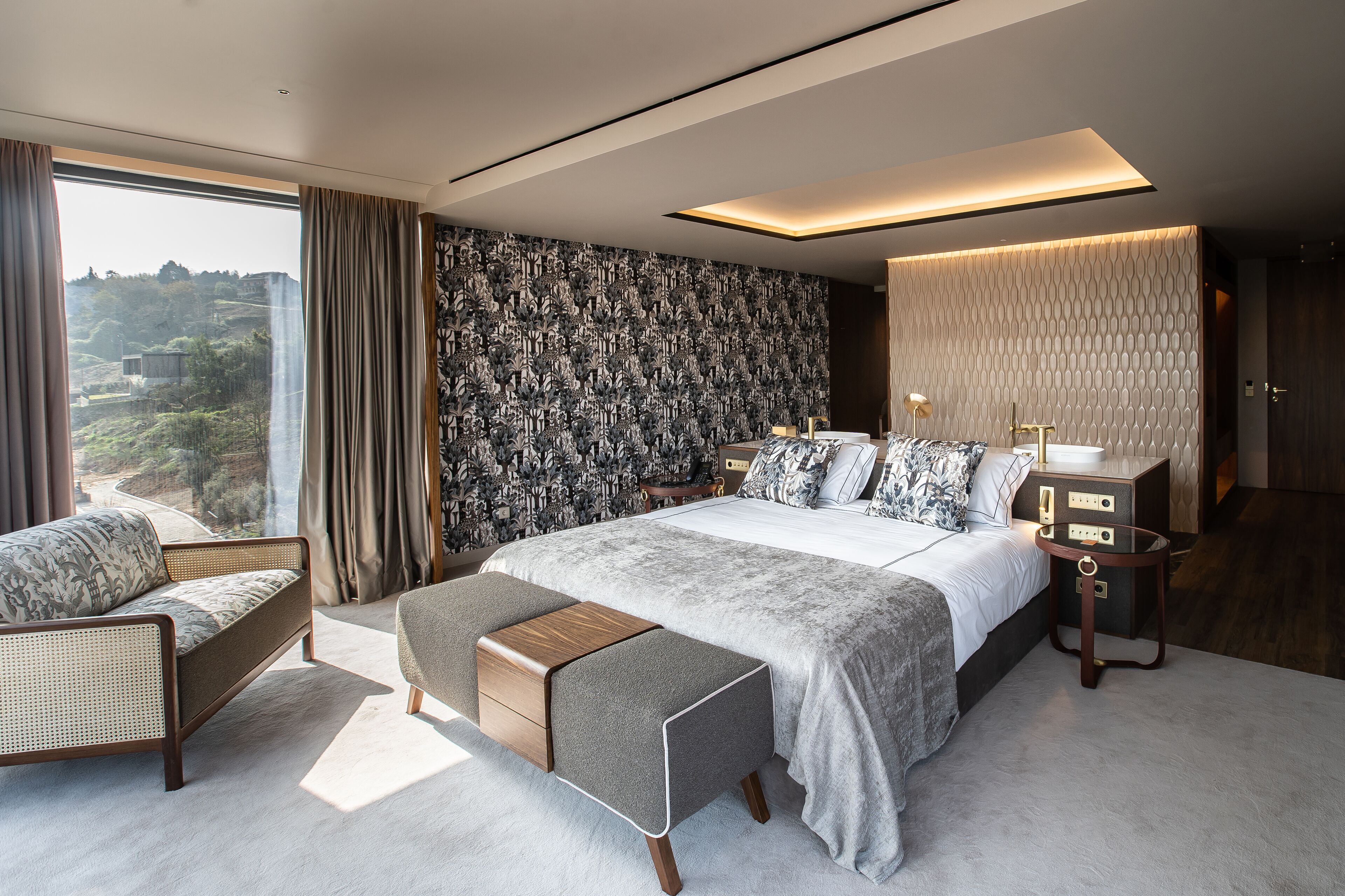 A modern bedroom with a large bed, a sofa, a bedside table, and a view of a mountainous landscape.