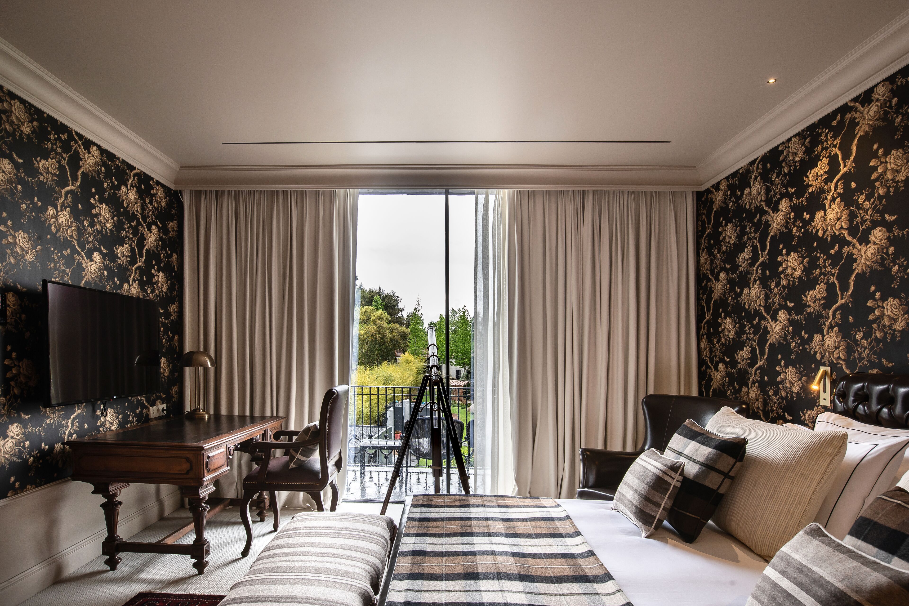 A bedroom with a bed, a desk, and a chair. There is a balcony with a view of greenery.