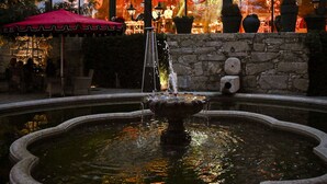 A building with large windows reflecting lights, a fountain, and outdoor seating with a red umbrella.