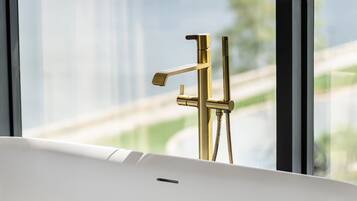 A bathroom with a bathtub, a gold faucet, and a view of a body of water through a window.