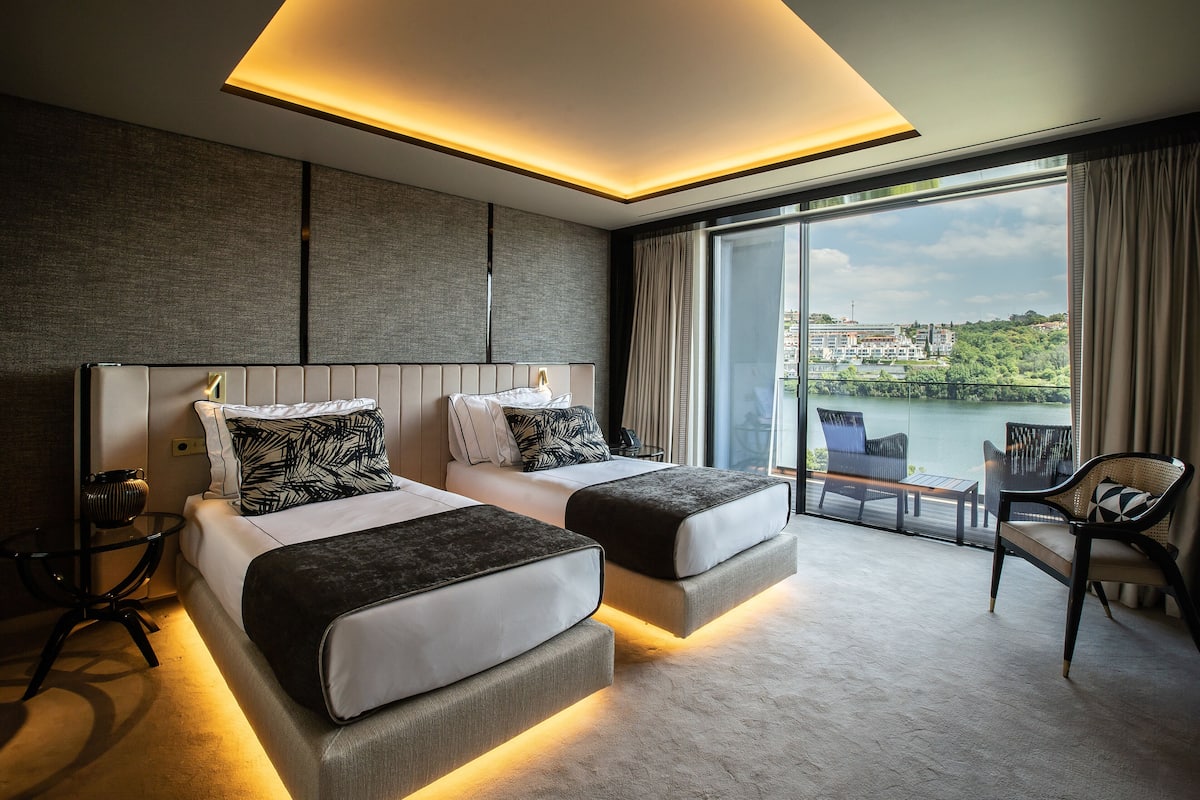 Une chambre d’hôtel moderne avec deux lits, un mur en verre et une vue sur un lac et des immeubles.