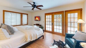 A bedroom with a bed, a ceiling fan, a fireplace, and large windows.