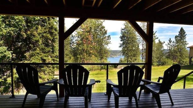 A covered deck with four Adirondack chairs facing a scenic view of a lake and forest.