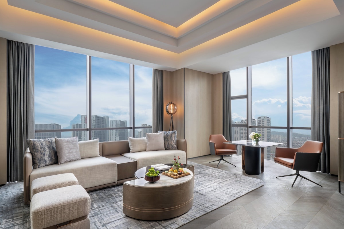 Une chambre d’hôtel moderne dotée d’une grande fenêtre, d’un canapé, d’une table basse ronde et d’un coin repas.
