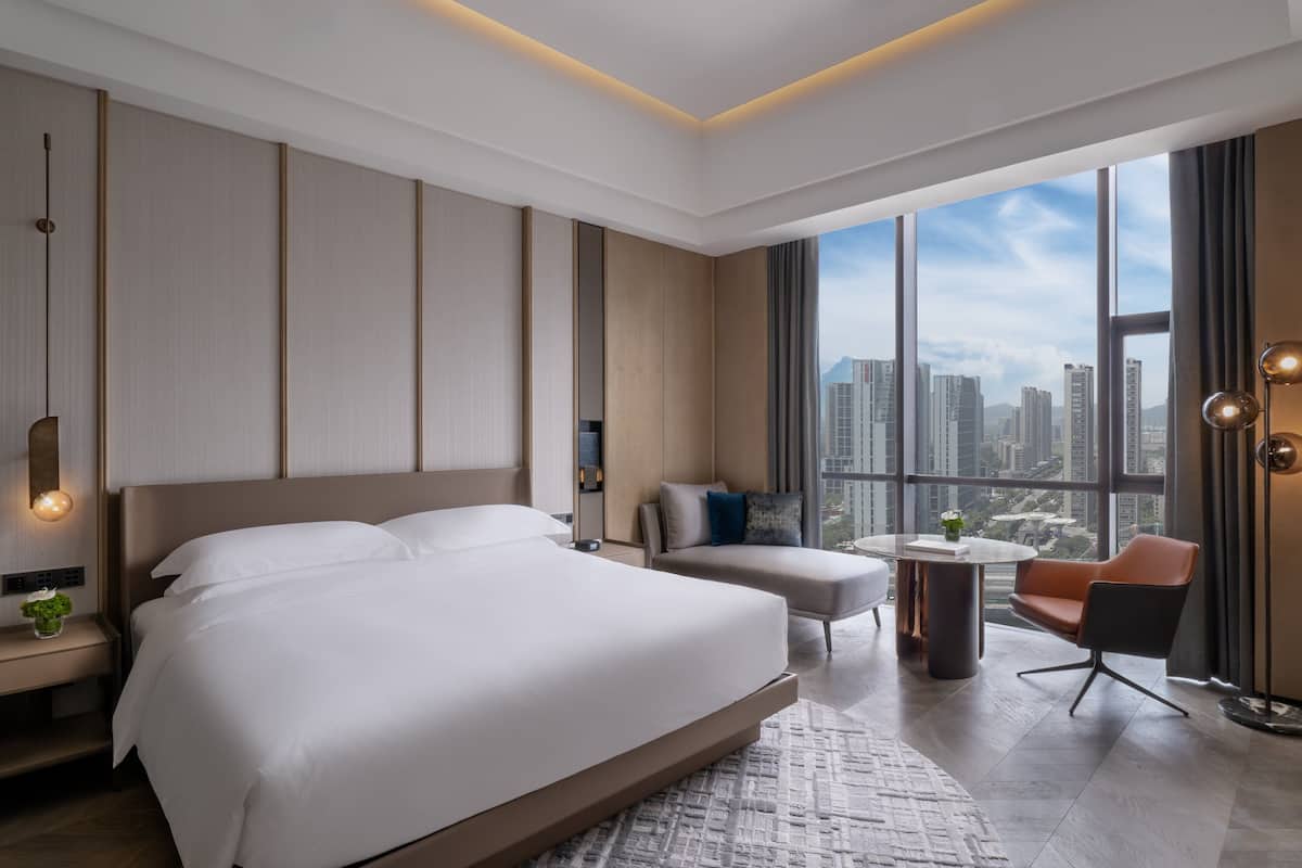 A modern hotel room with a large bed, a sofa, a small table, and a view of the cityscape through large windows.