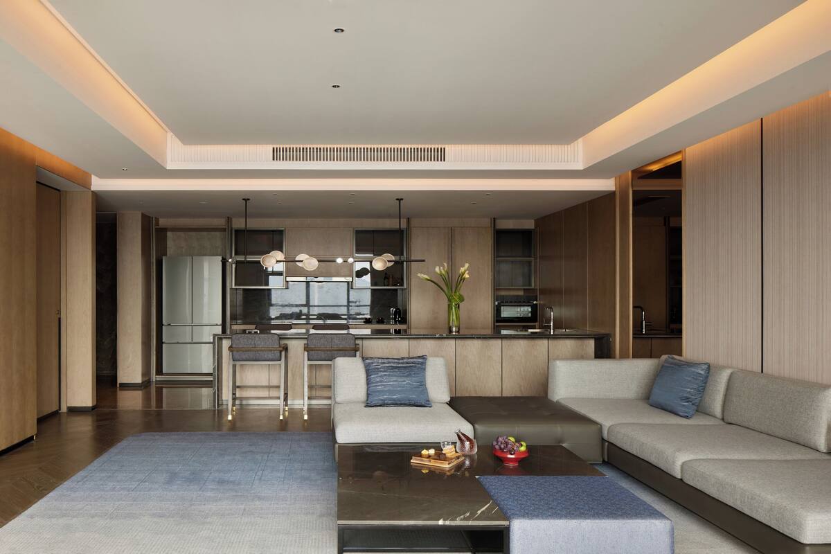A modern living room with a sofa, a coffee table, and a TV. There is a kitchen in the background with a dining area.