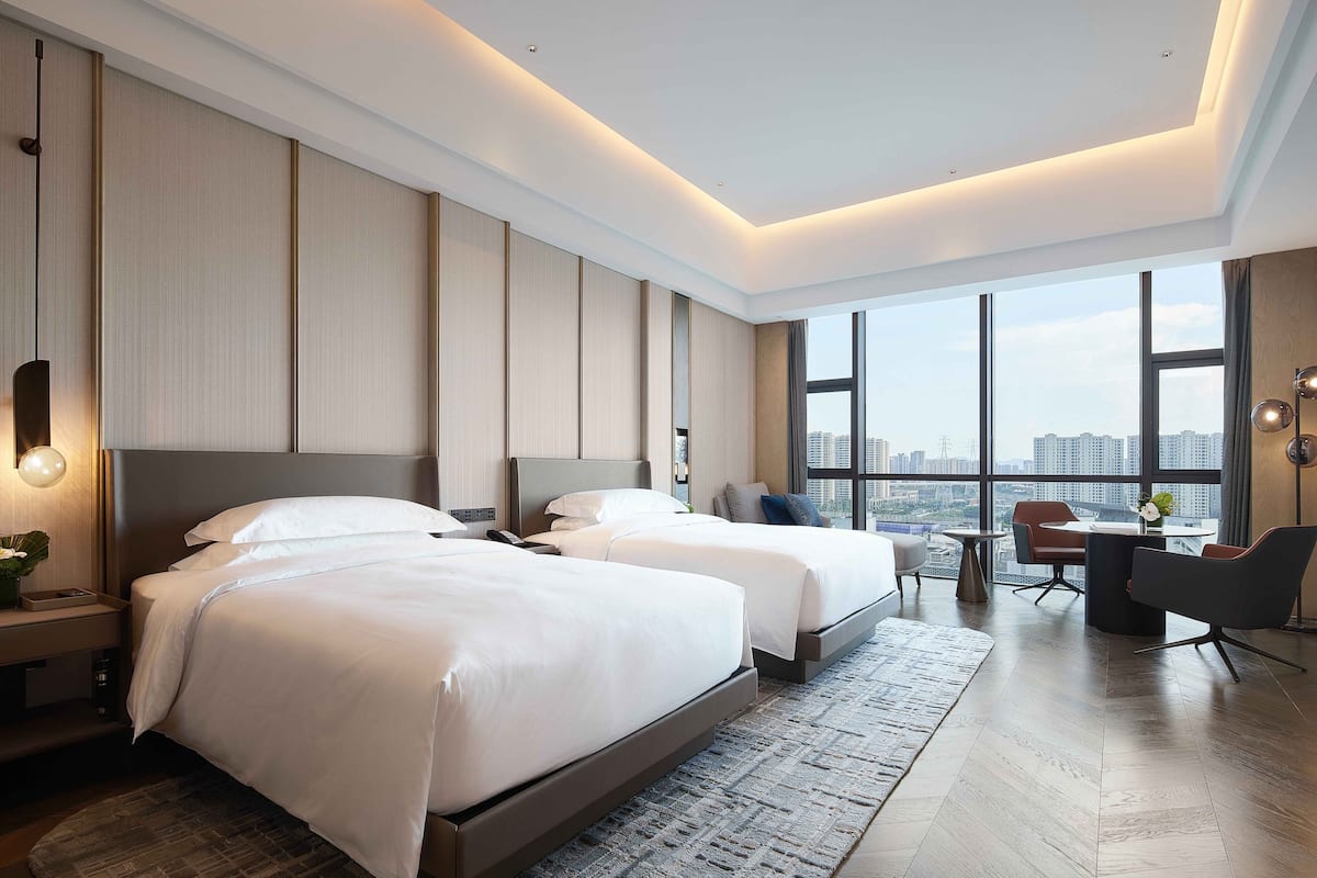 A modern hotel room with two beds, a desk, and a view of the city.