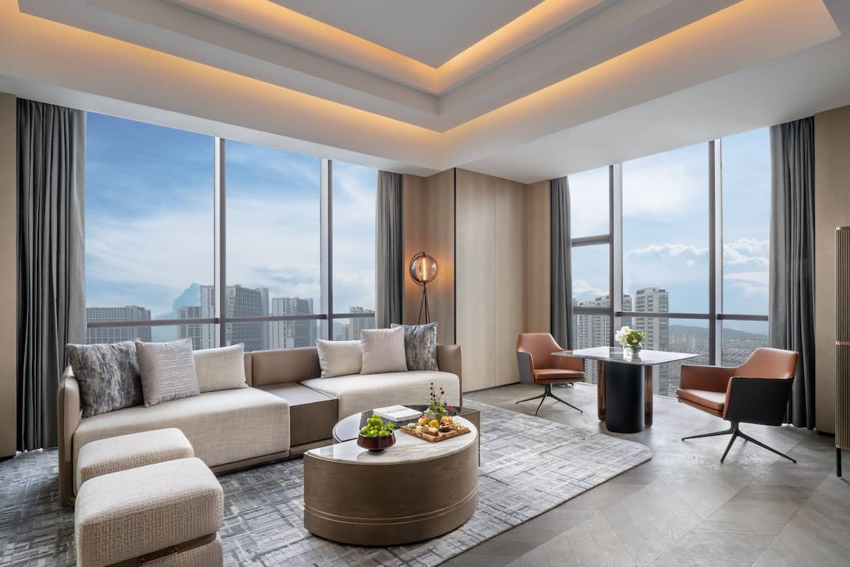 A modern hotel room with a large window, a sofa, a round coffee table, and a dining area.