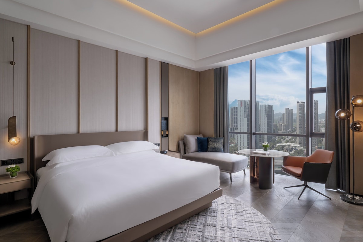 Une chambre d’hôtel moderne, dotée d’un grand lit, d’un canapé, d’une petite table et offrant une vue sur le paysage urbain grâce à de grandes fenêtres.
