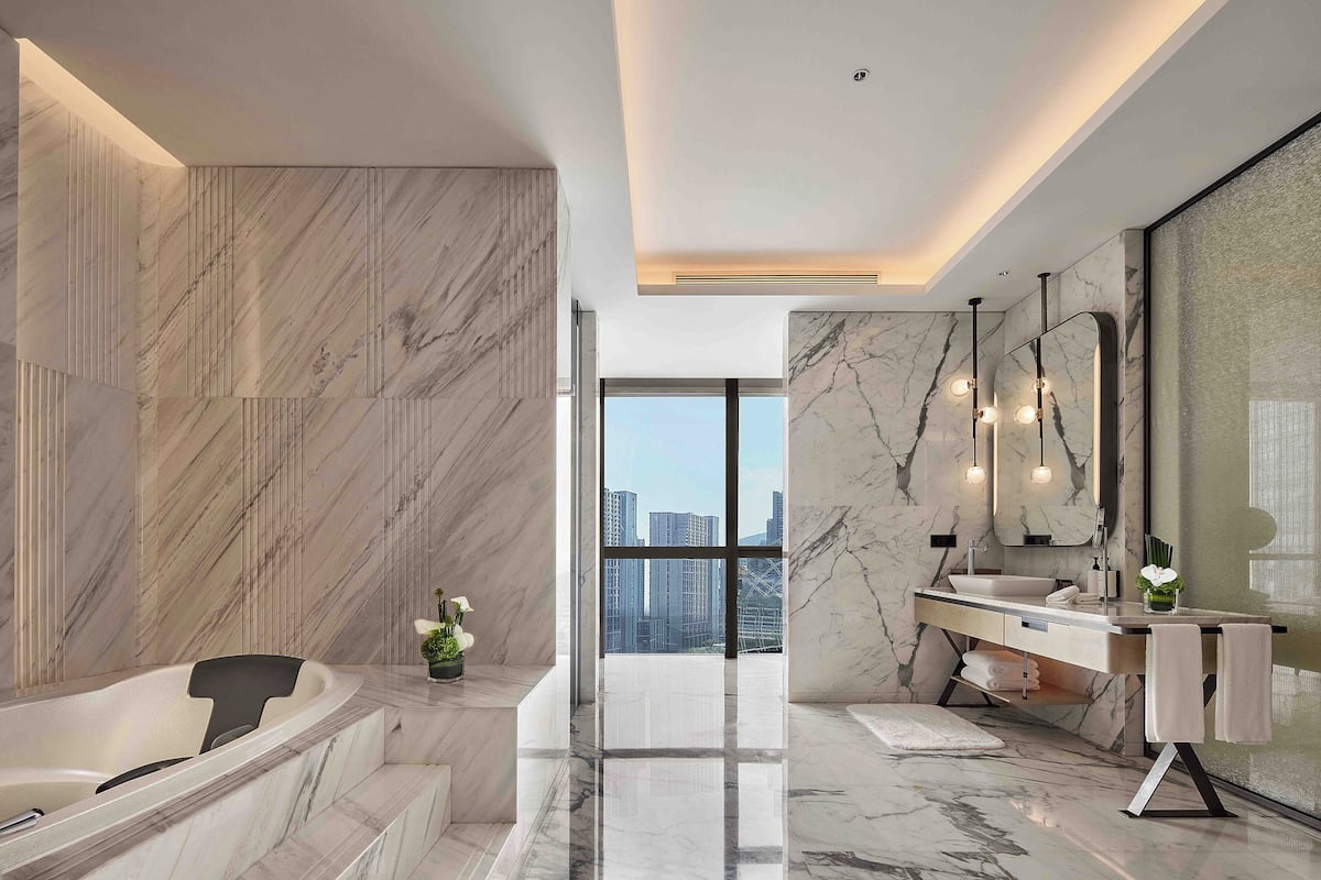 A modern bathroom with a marble floor, a bathtub, a vanity with a large mirror, and a view of the city through large windows.