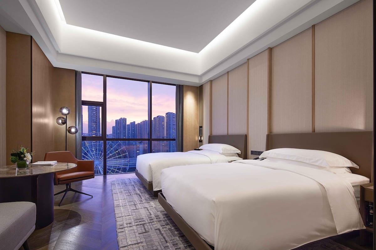 A hotel room with two beds, a desk, and a chair. The room has a large window with a city view.