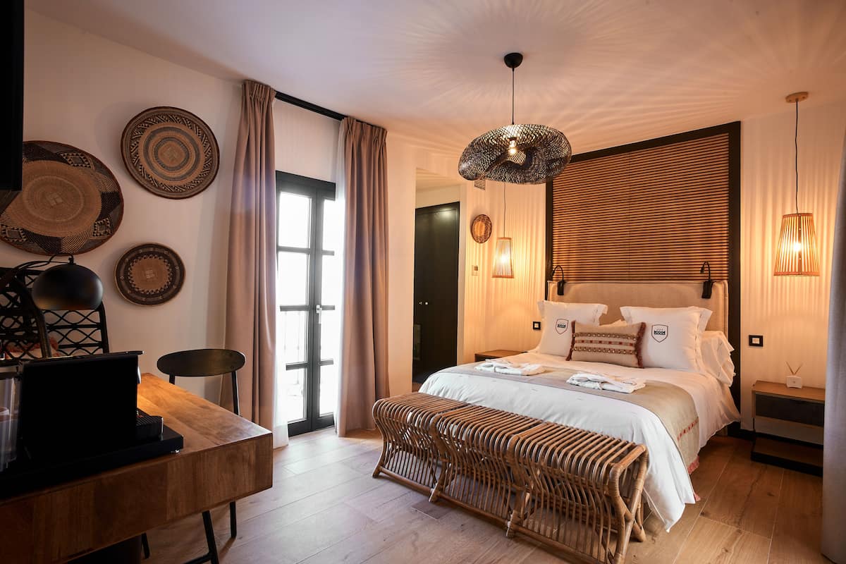 A modern bedroom with a large bed, a desk with a laptop, and a wicker bench.