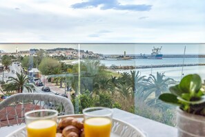 A balcony with a table set for breakfast, overlooking a coastal town with buildings, a beach, and a lighthouse.