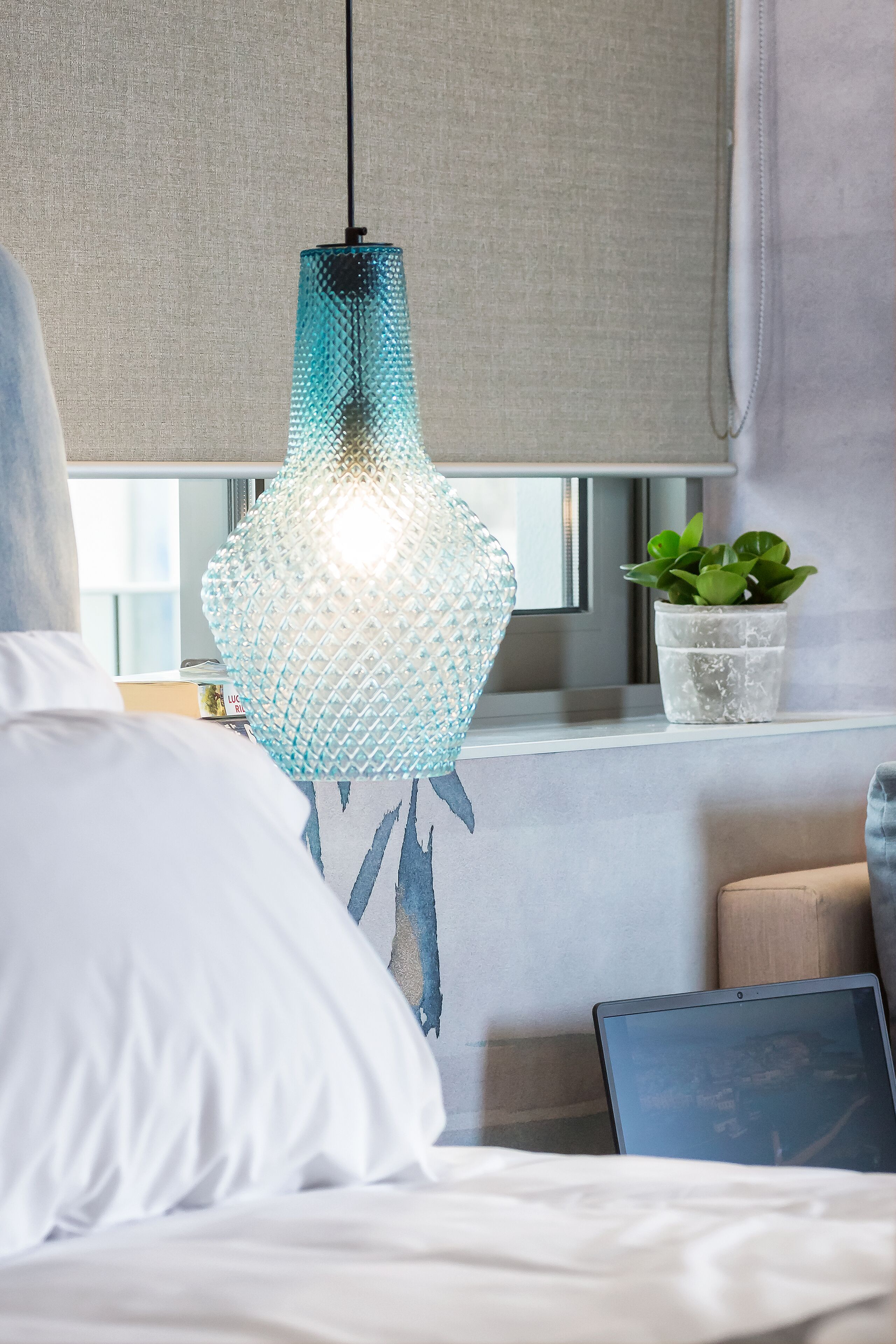 A bed with white bedding, a laptop on the side, a hanging light fixture, a plant, and a window with curtains.