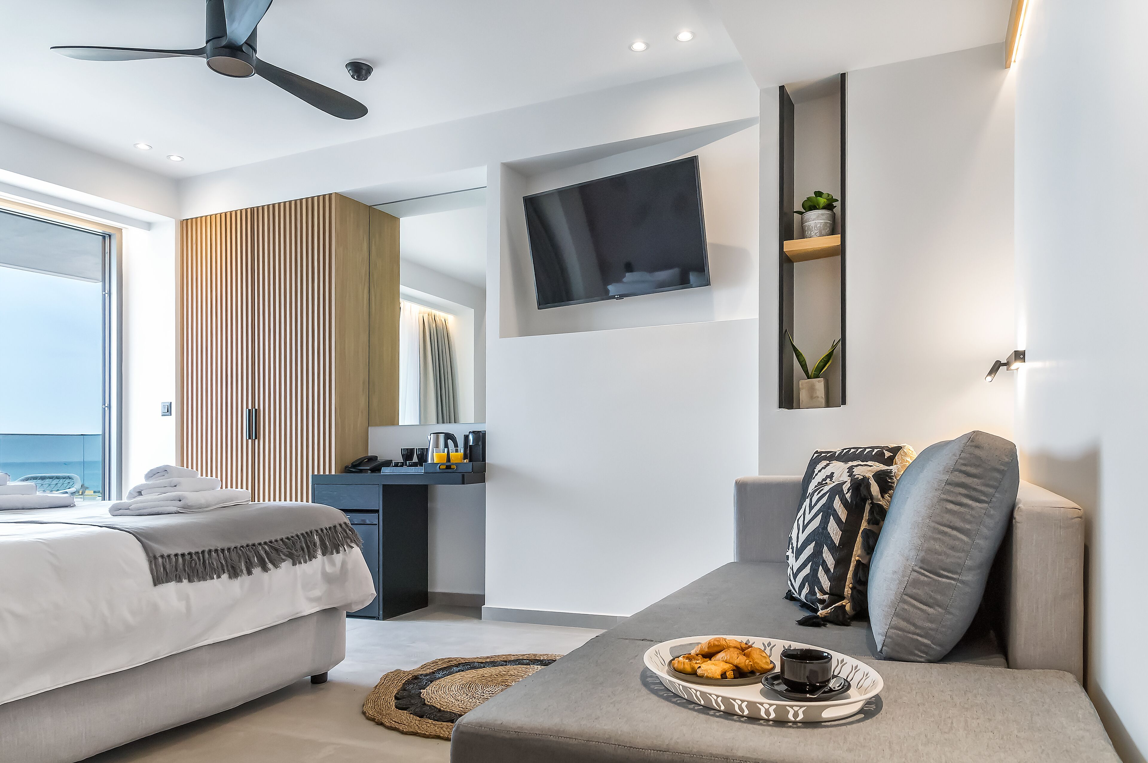 Una habitación de hotel moderna con una cama, un sofá, un televisor instalado en la pared y un pequeño escritorio con una planta.