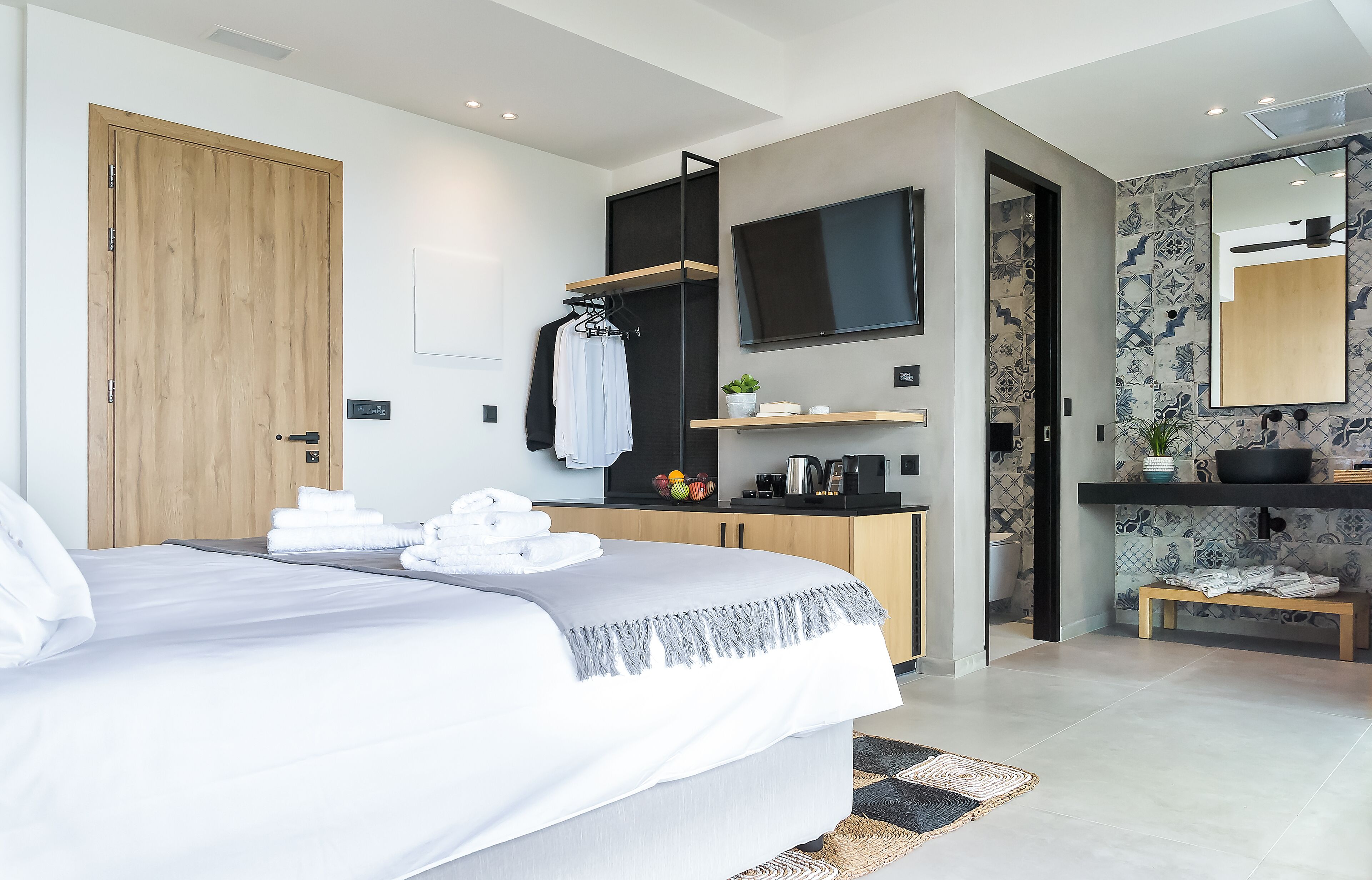 Un dormitorio moderno con cama, puerta de madera, televisor, baño con lavabo y espejo, y una planta pequeña.
