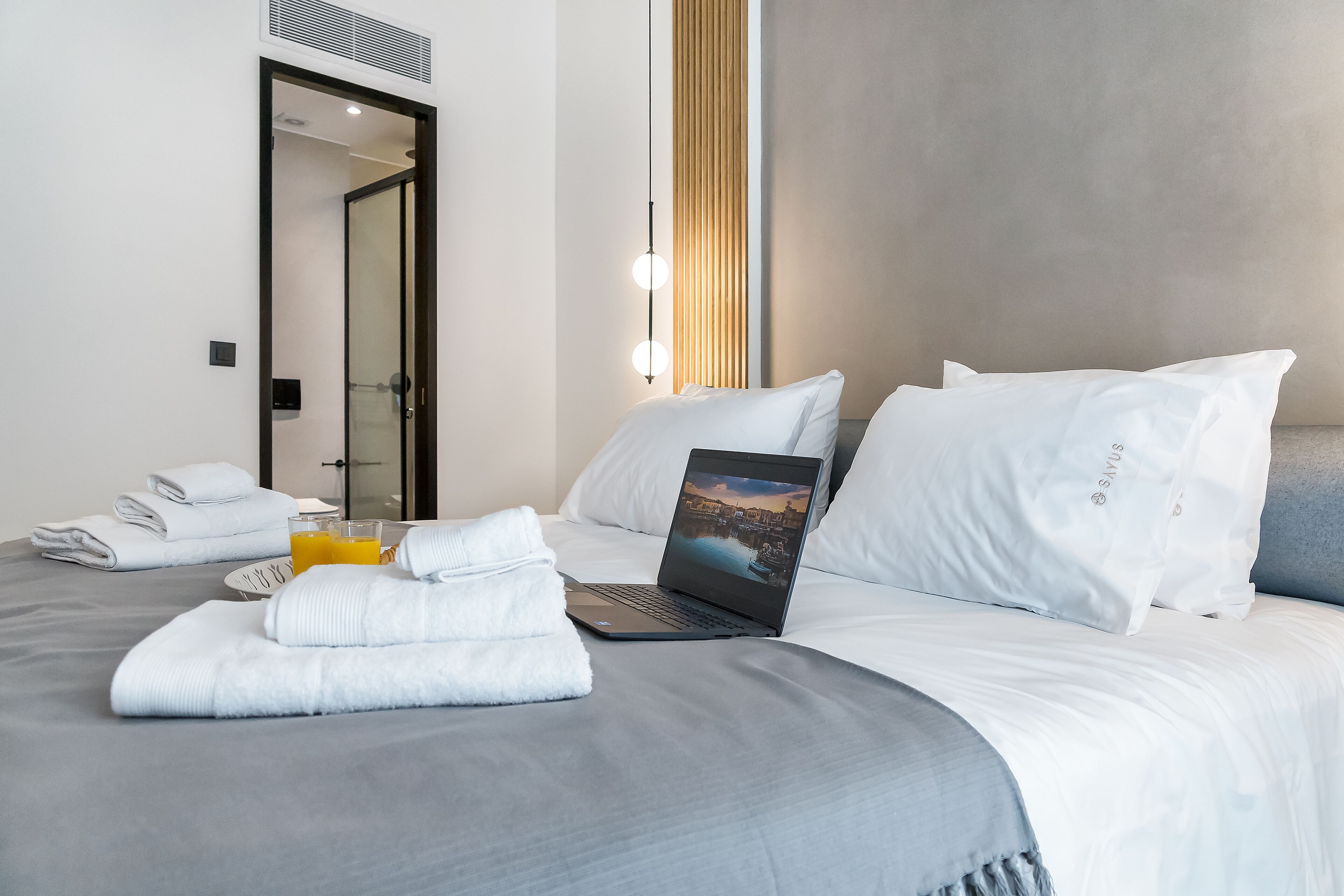 Una cama bien tendida con un ordenador portátil, un vaso de zumo de naranja y toallas dobladas.