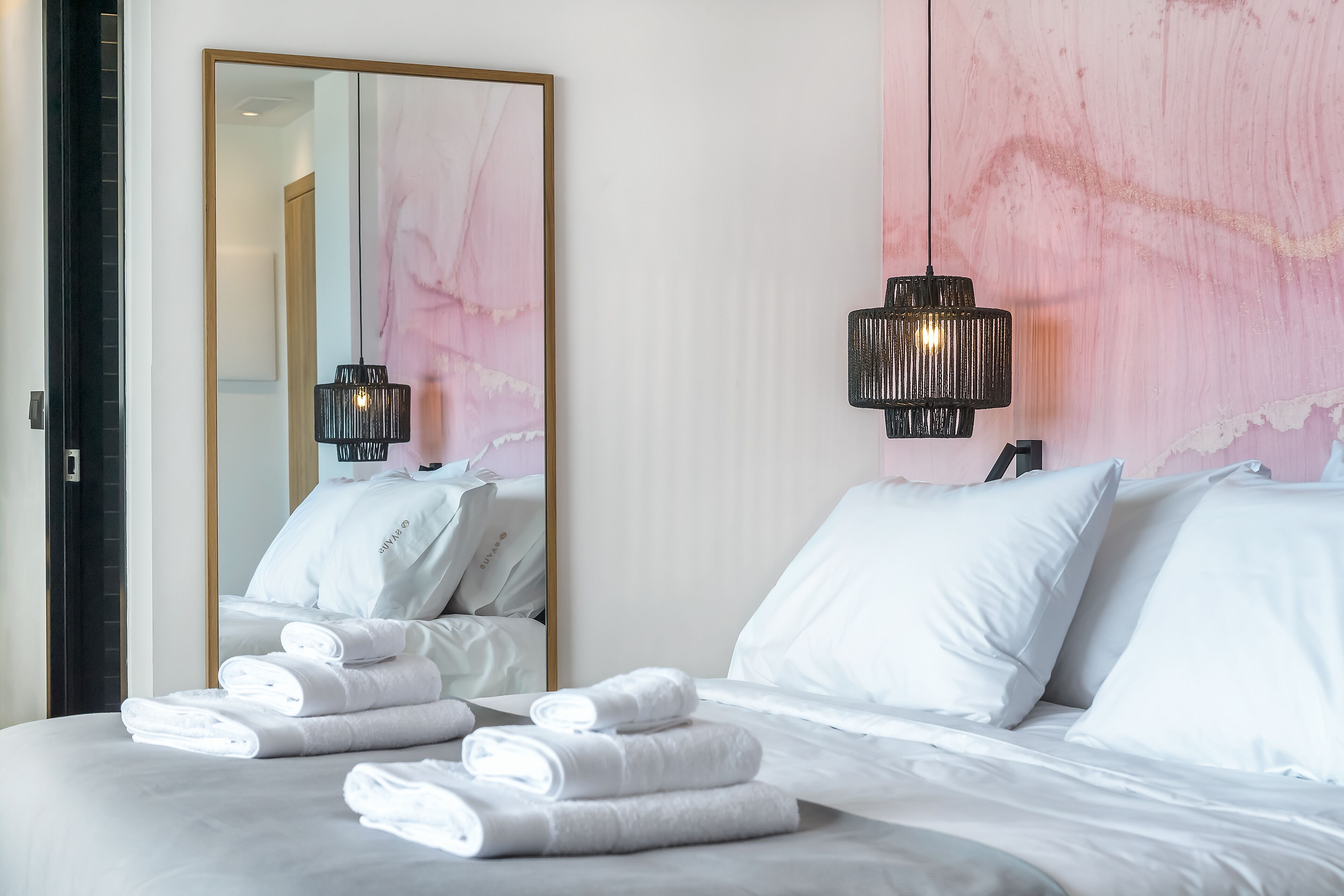 A hotel room with a bed, white towels, a large mirror, and a pink abstract wall.