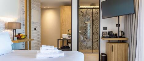 Habitación de hotel con cama, televisor, escritorio pequeño con lámpara, frutero y baño con ducha e inodoro.