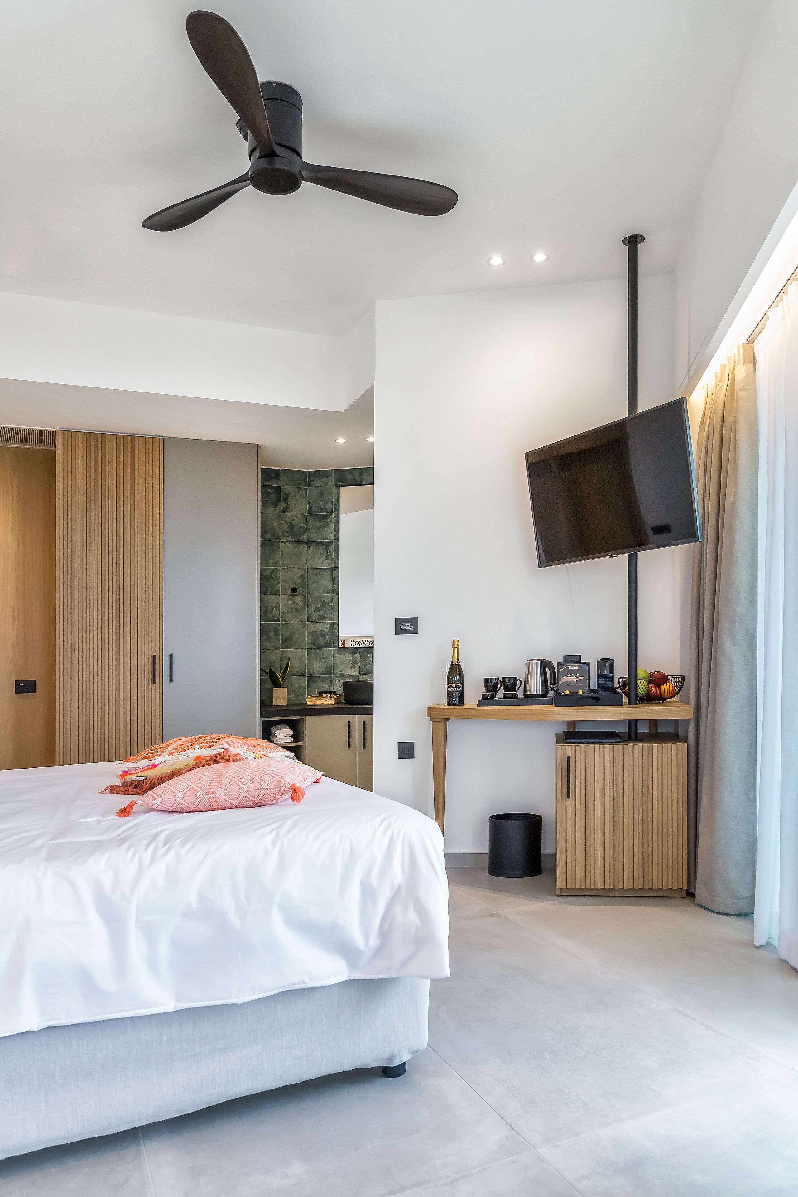 Una habitación de hotel moderna con cama, escritorio con televisor de pantalla plana, ventilador de techo y una pequeña cocina.