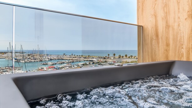 Une salle de bain moderne avec une baignoire entourée de parois de verre, donnant sur un port de plaisance et la mer.