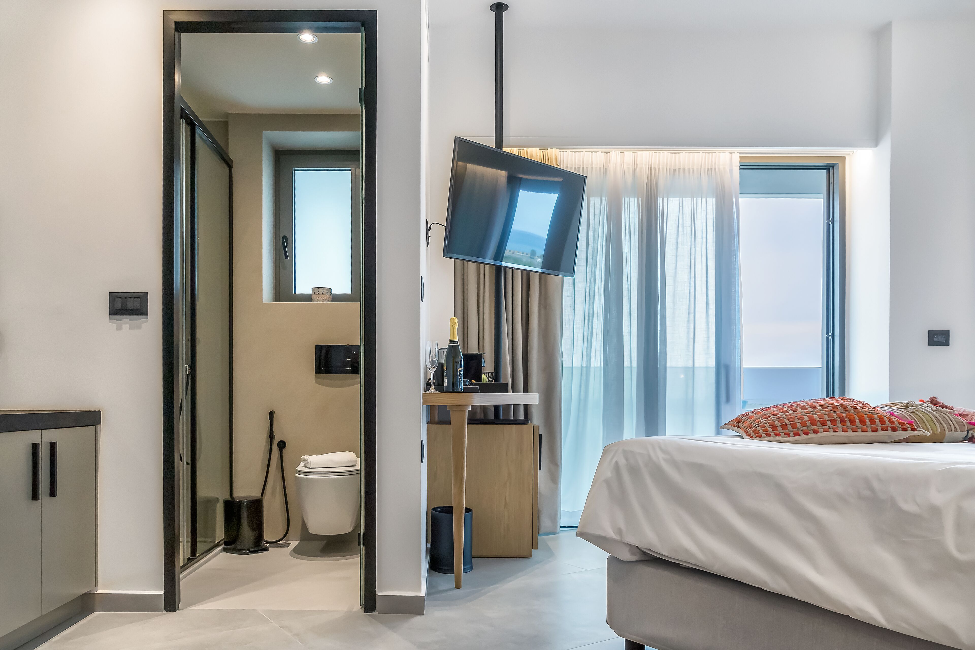 Une chambre d’hôtel moderne, équipée d’un lit, d’un bureau, d’une télévision et d’une salle de bain visible à travers une porte ouverte.