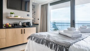 Une chambre d’hôtel moderne dotée d’un grand lit, d’un téléviseur à écran plat fixé au mur, d’un petit coin cuisine avec une machine à café et d’un balcon offrant une vue sur la mer.
