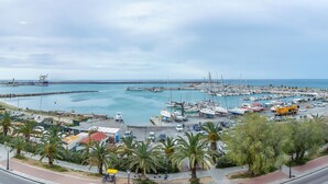 Un port de plaisance sur la côte, avec des bateaux amarrés, une route où circulent des véhicules et une promenade piétonne bordée de palmiers.