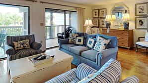 Ein Wohnzimmer mit einem blauen Sofa, gestreiften Sesseln, einem weißen Couchtisch und einem Balkon mit Korbmöbeln.