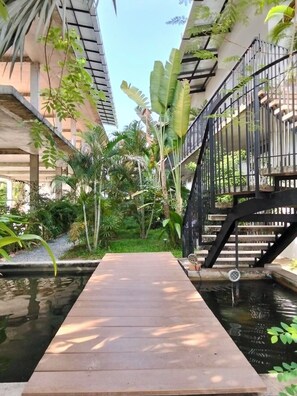 Eine Holzterrasse über einem Wasserspiel, umgeben von üppiger Bepflanzung und einer schwarzen Metalltreppe.