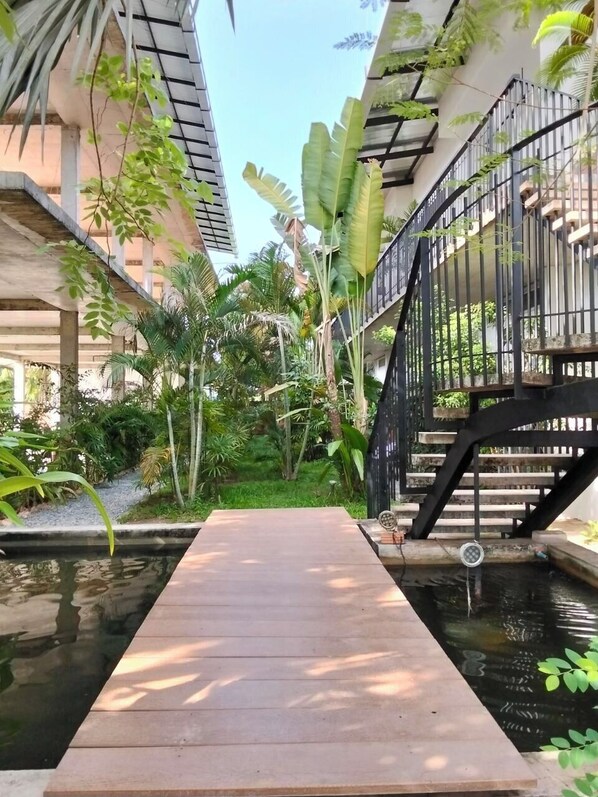 A wooden deck over a water feature, surrounded by lush greenery and a black metal staircase.