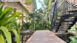 Eine Holzterrasse führt zu einem Wasserspiel, umgeben von üppiger Vegetation und einer schwarzen Metalltreppe.