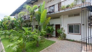 Ein mehrstöckiges Gebäude mit einem Balkon im zweiten Stock, umgeben von üppiger Vegetation und einem Kiesweg.