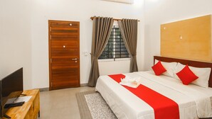 Ein Hotelzimmer mit Holztür, einem Bett mit rot-weißer Bettwäsche, einem Schreibtisch mit Computer und einem Fenster mit Vorhängen.