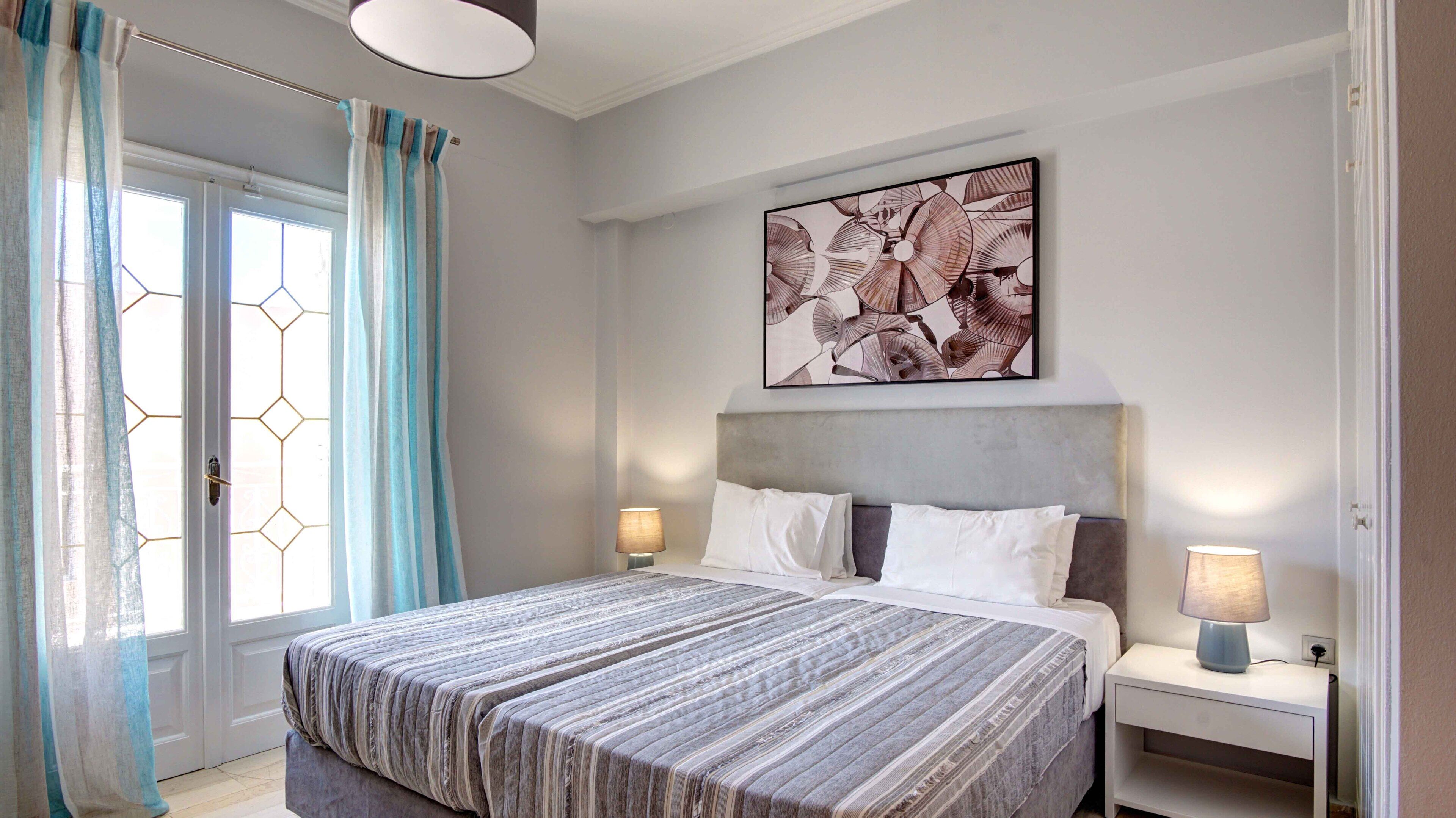 Une chambre à coucher avec un grand lit, deux tables de chevet avec des lampes, une fenêtre avec des rideaux et une œuvre d’art accrochée au mur.