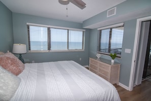 Un dormitorio con dos ventanas, una cama con colcha blanca, un tocador y una lámpara.