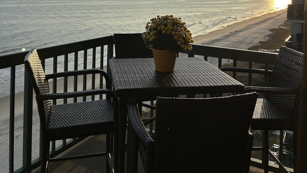 A balcony with wicker furniture and a potted plant, overlooking a beach at sunset.