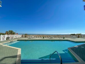 Una piscina azul cristalina con cubierta de madera, sillas de descanso y vistas al océano.