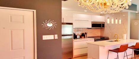 Una cocina moderna con gabinetes blancos, una isla central con taburetes de bar, una araña y un pequeño adorno de sol en la pared.
