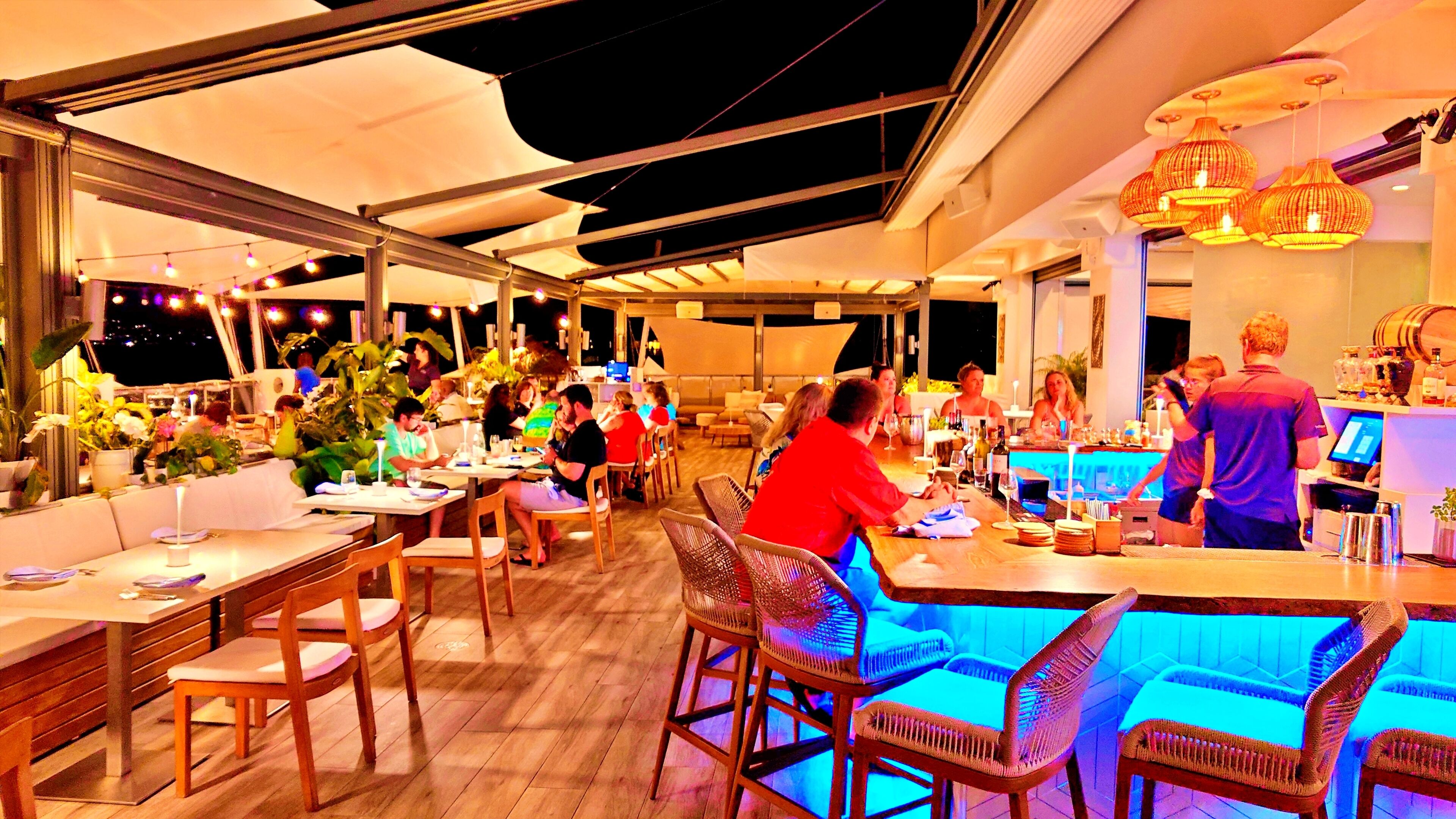 Ein Außenbereich mit Essbereich, Tischen und Stühlen, eine Bar mit Theke und Hockern sowie ein großer Sonnenschirm, der Schatten spendet.
