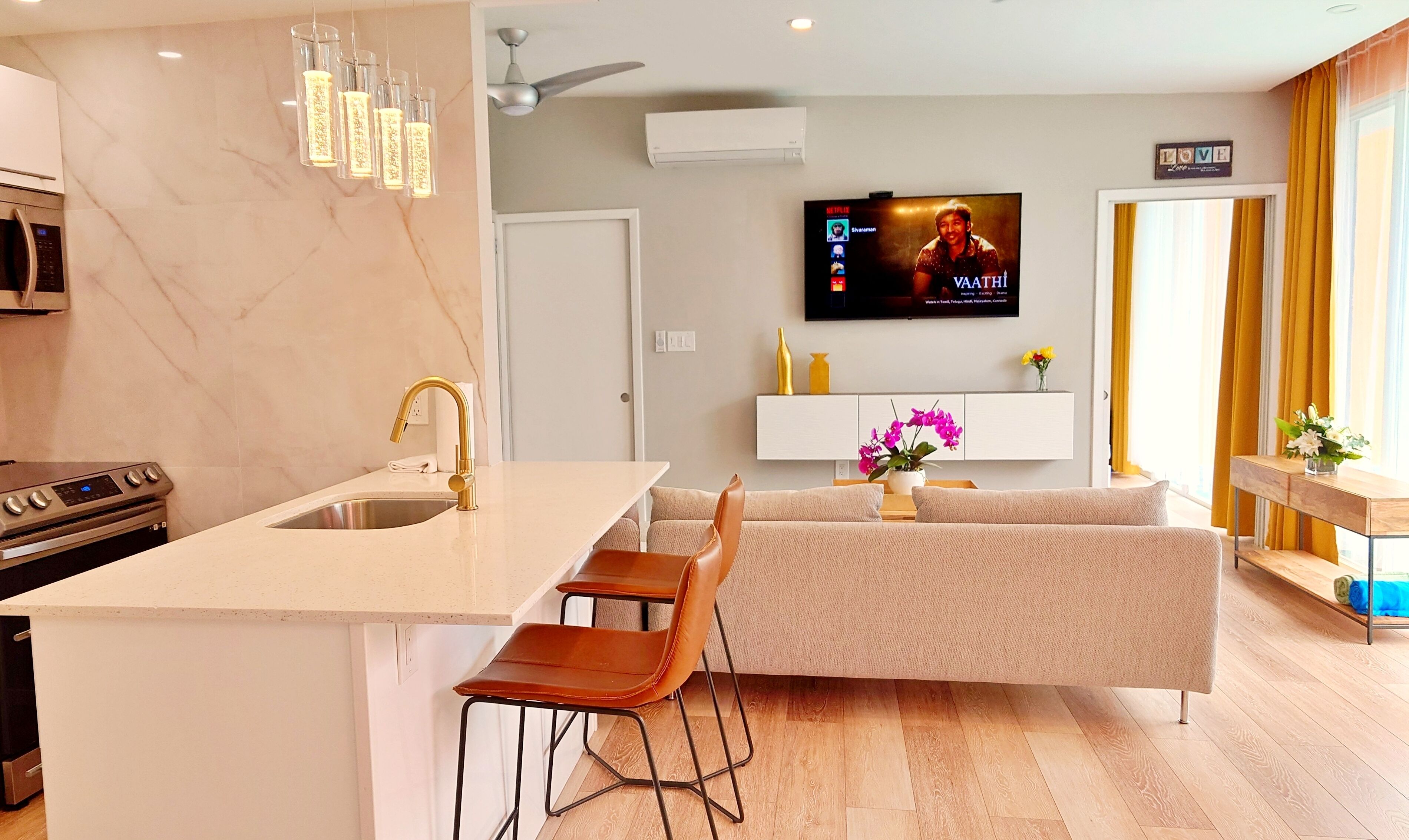 A modern kitchen with a central island, a dining area with a sofa and chairs, a flat-screen TV mounted on the wall, and a window with curtains.