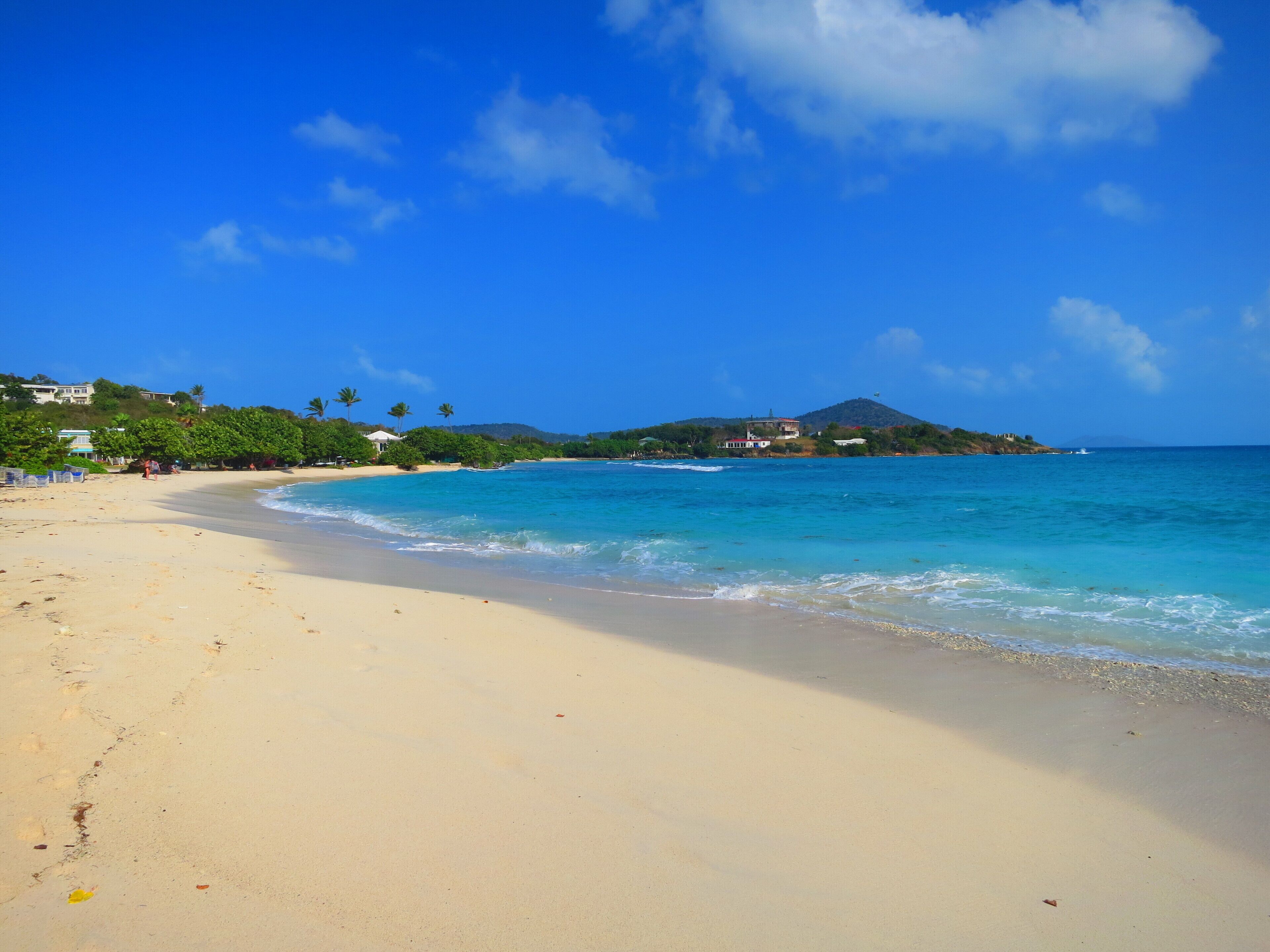 A sandy beach with clear blue waters and a mountain in the background.