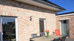 Een terras buiten met een tafel en stoelen, een vaas met bloemen en een bakstenen muur.