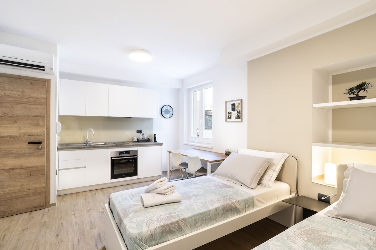 A modern, well-lit room with a bed, bedside table, and a small desk with a chair. A kitchen area with white cabinets and a sink is visible in the background.