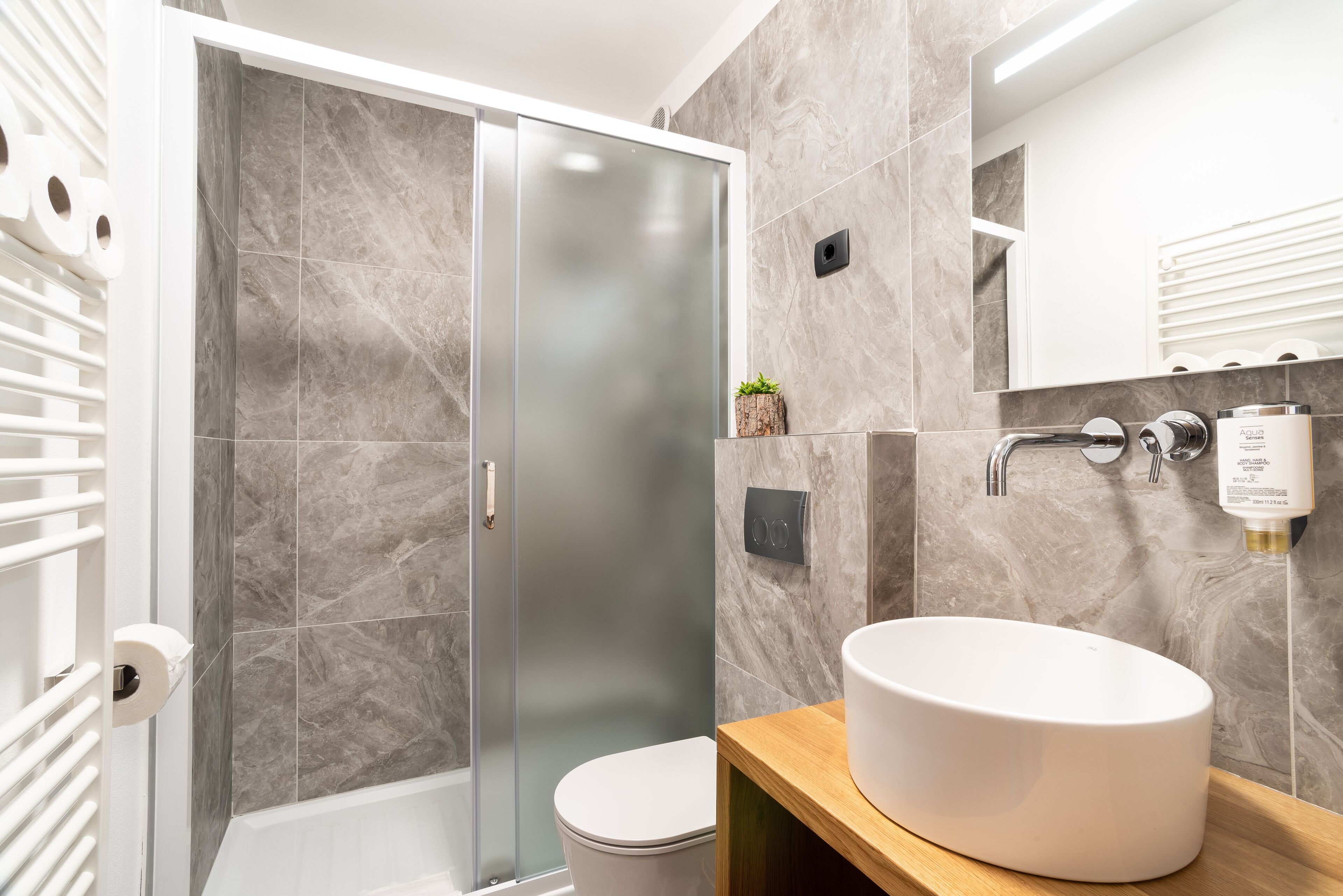 Un baño moderno con ducha envuelta en vidrio, inodoro blanco, tocador de madera con lavamanos blanco y pared con azulejos de mármol.