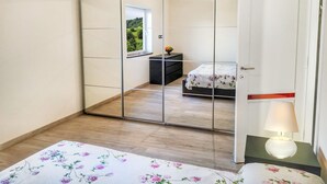 A bedroom with a mirrored wardrobe, a bed with a floral bedspread, a nightstand with a lamp, and a view of a garden through a window. - Rosalba, Private solar heated pool, walk restaurants, romantic, mountain views.  (Corfino)