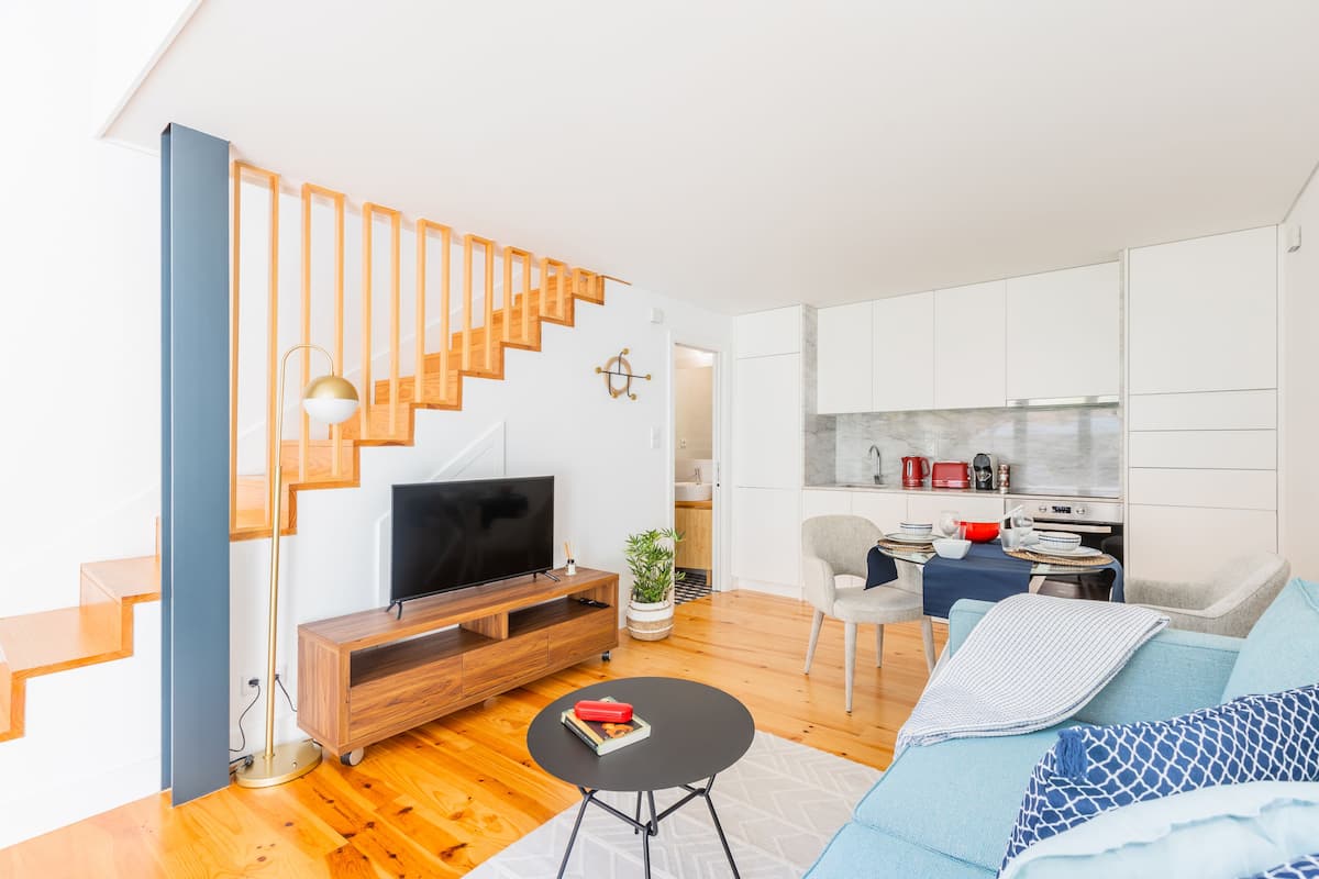 A modern living room with a wooden staircase, a flat-screen TV, a coffee table, and a sofa with patterned cushions.