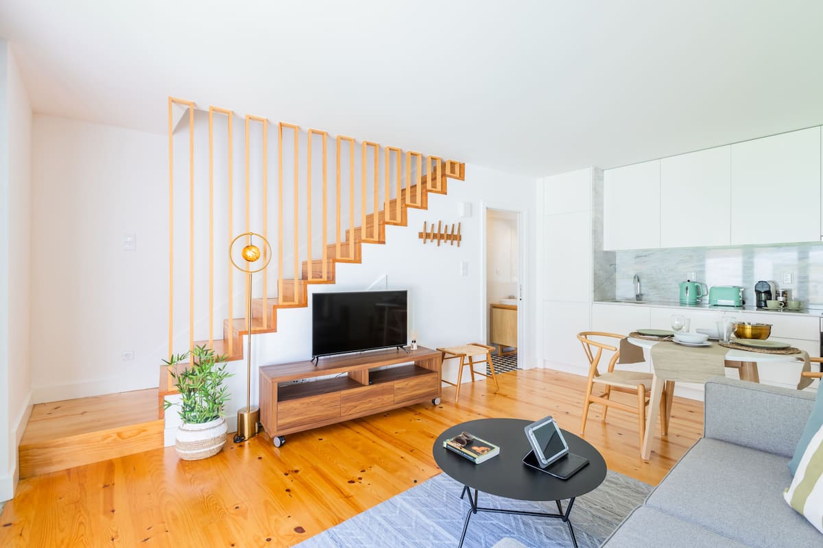A modern living room with a wooden staircase, a flat-screen TV, a dining area with a kitchen in the background, and a potted plant.